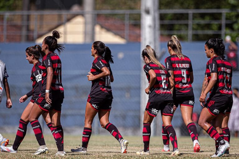 Jogadoras de futebol usando uniformes preto e vermelho listrados caminham pelo campo durante uma partida. Supercopa Feminina.